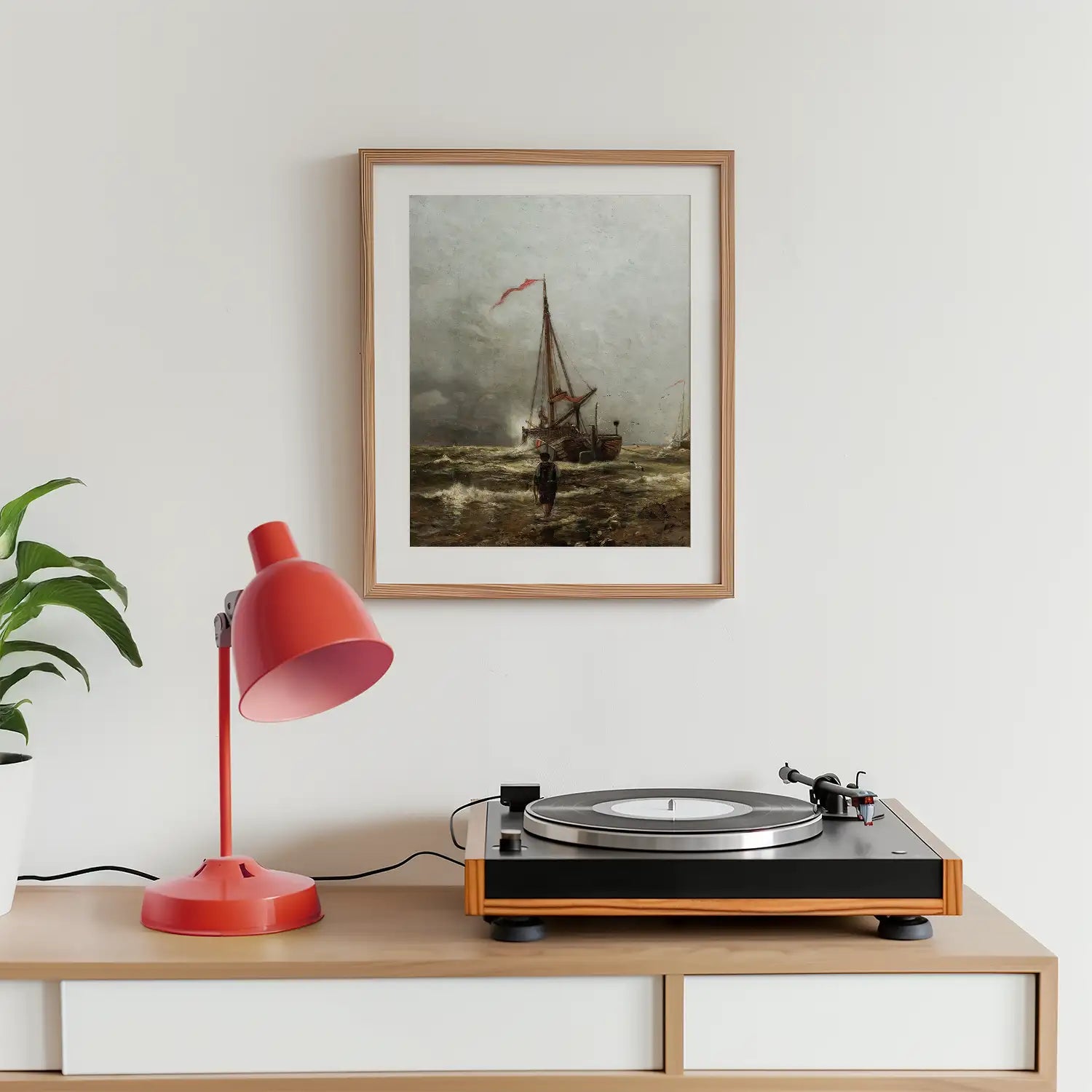 Framed painting of a sailboat at sea hanging on a wall above a record player and a red desk lamp, with a green plant in the background.