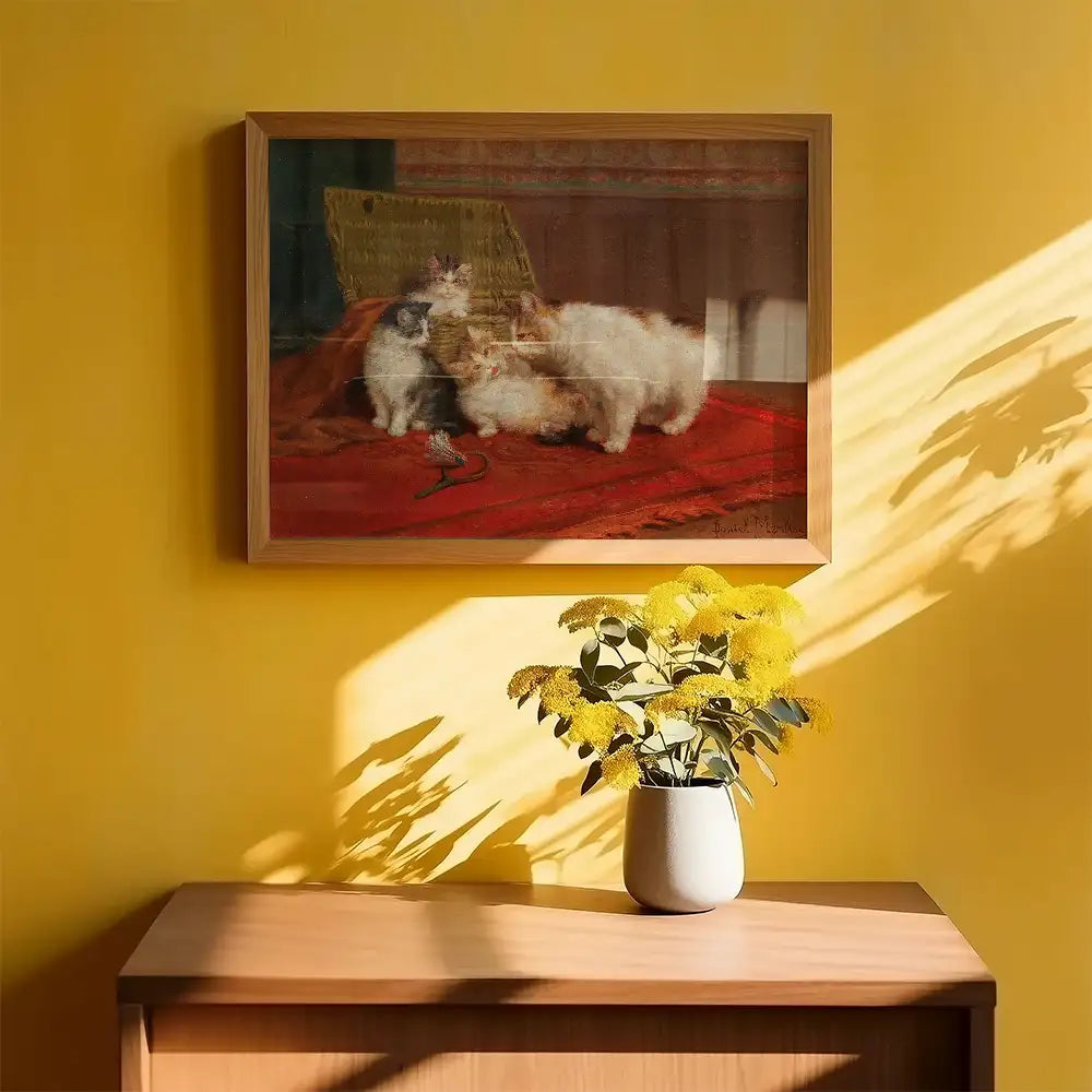 Painting of two cats on a red cloth, framed and hung on a yellow wall with a vase of yellow flowers on a wooden table.