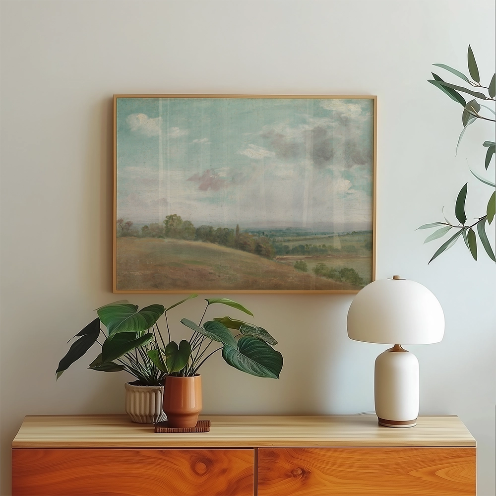 Landscape painting in a wooden frame displayed on a light wood sideboard with a potted plant and a modern lamp.