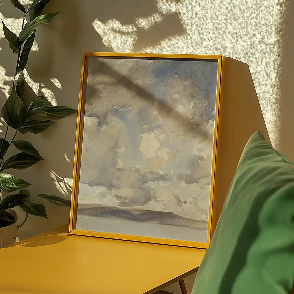 Framed watercolor painting of a cloudy sky displayed on a yellow table, surrounded by greenery and soft furnishings.