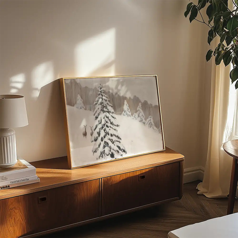 Framed painting of a snowy landscape featuring a snow-covered tree and deer, displayed on a wooden surface with a lamp and books nearby.