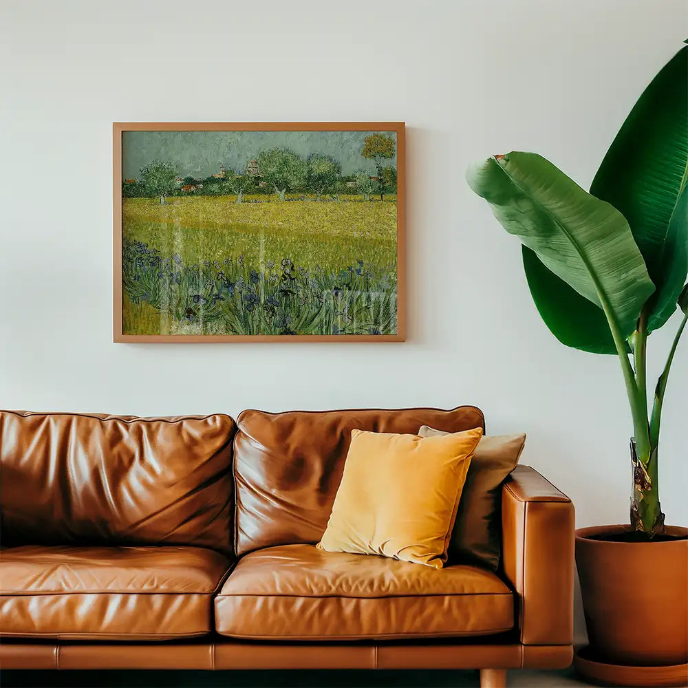 Framed painting of a vibrant yellow field with trees and a blue sky, displayed above a brown leather sofa with a yellow pillow and a green plant nearby.