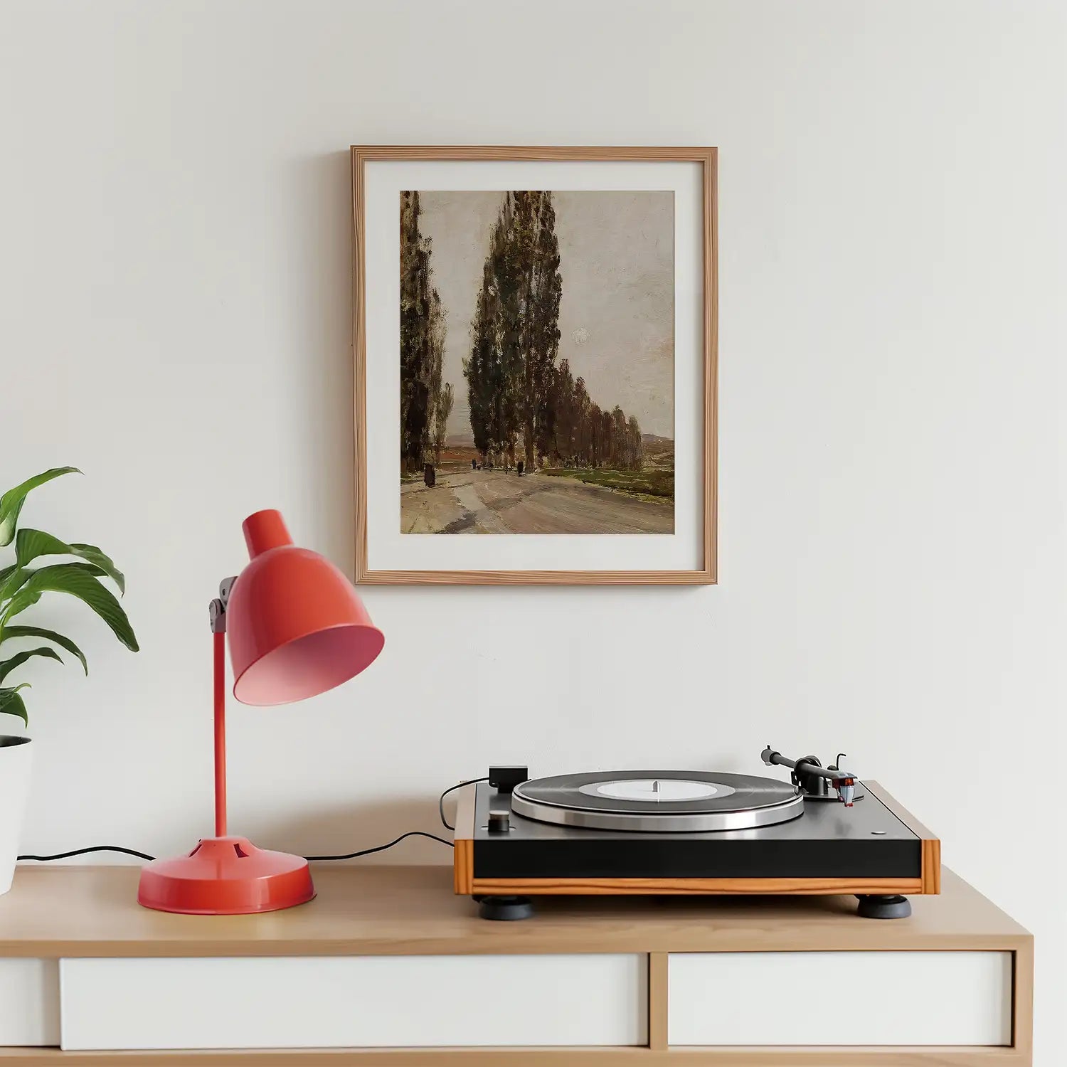 Framed painting of tall trees along a road, displayed above a red desk lamp and a record player on a wooden surface.