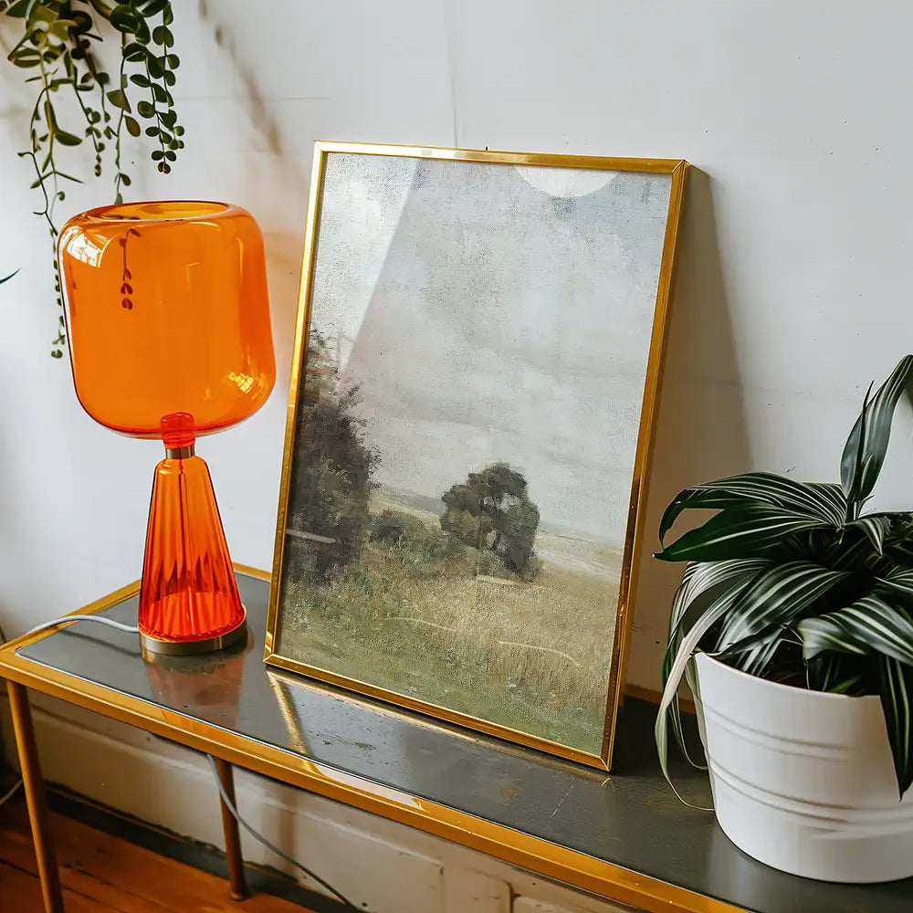 Painting in a gold frame displayed on a table next to an orange lamp and a potted plant.