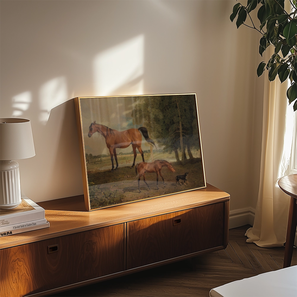 Framed painting of two horses and a dog in a pastoral setting, displayed on a wooden side table with a lamp and plants in a sunlit room.