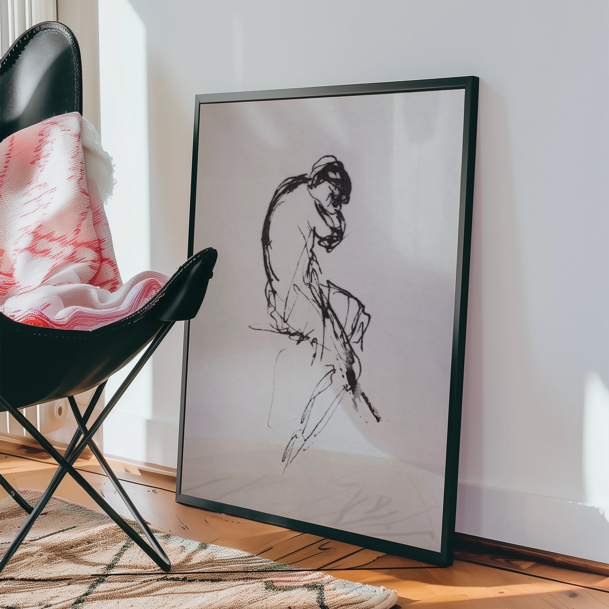 A black-framed painting of a seated figure in charcoal sketch style, leaning forward, resting their head on their hand, placed against a white wall next to a black chair and a patterned rug.