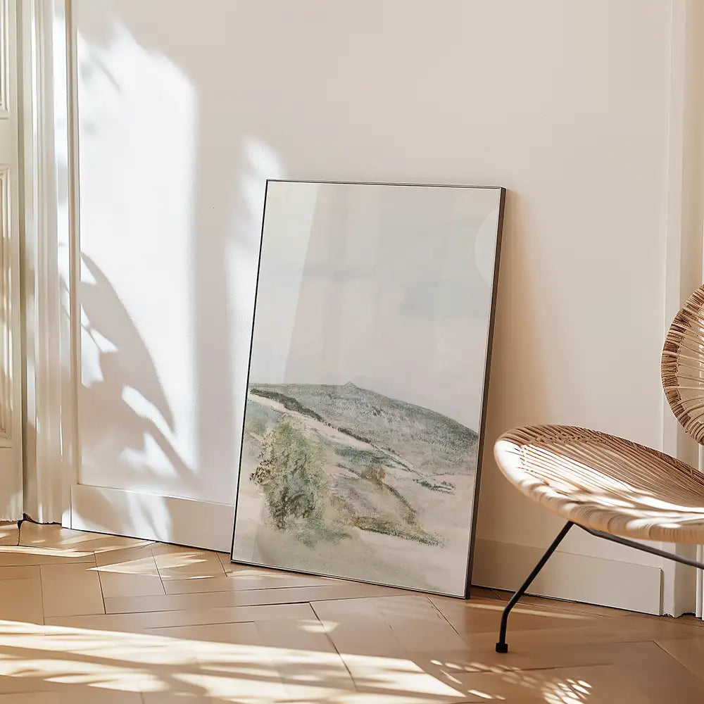 Landscape painting in a modern frame, leaning against a white wall beside a rattan chair.