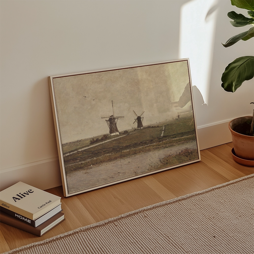 Framed painting of a landscape with windmills, positioned on a wooden floor beside stacked books and a potted plant.