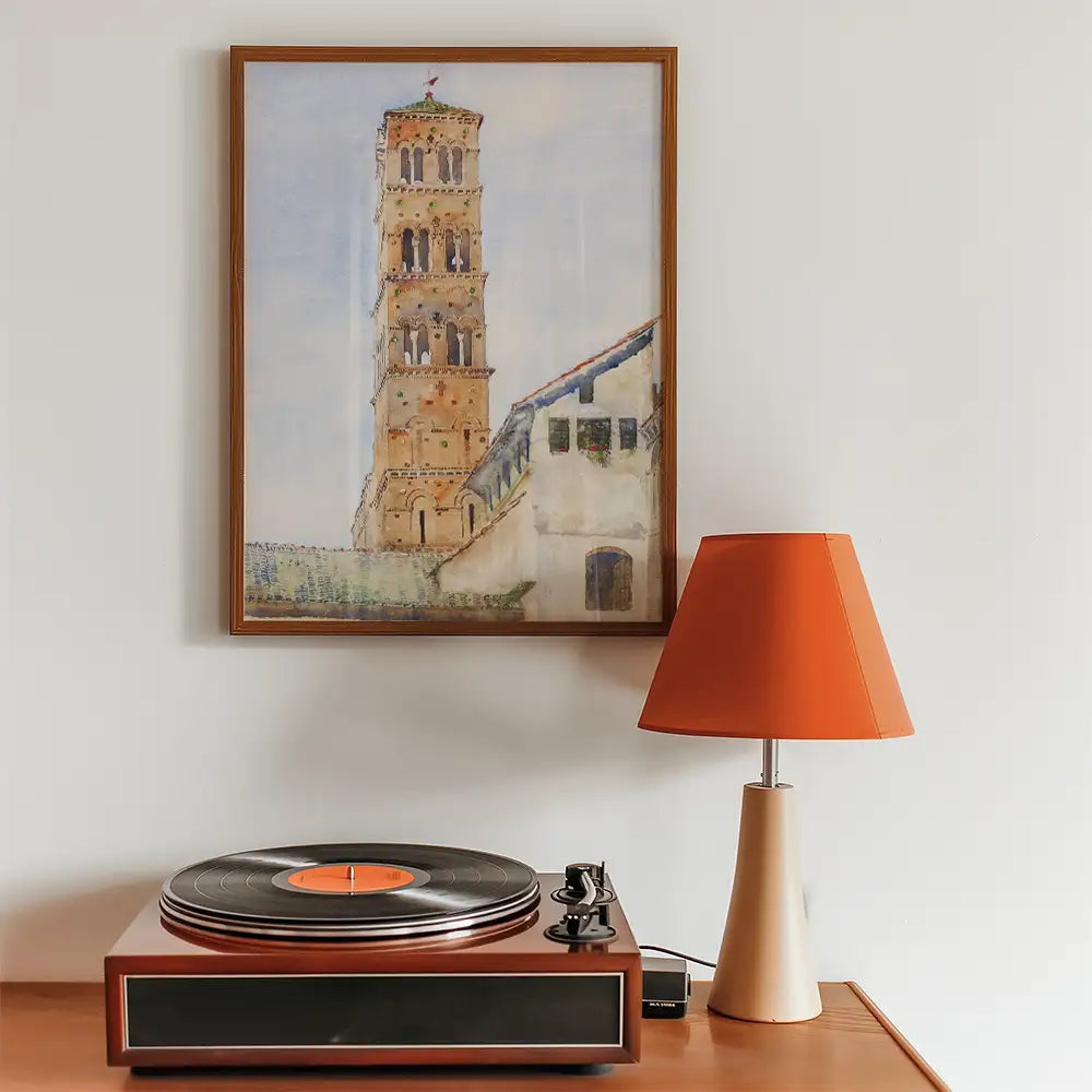Painting of a historical tower framed on a wall above a wooden table with a vintage record player and an orange lamp.