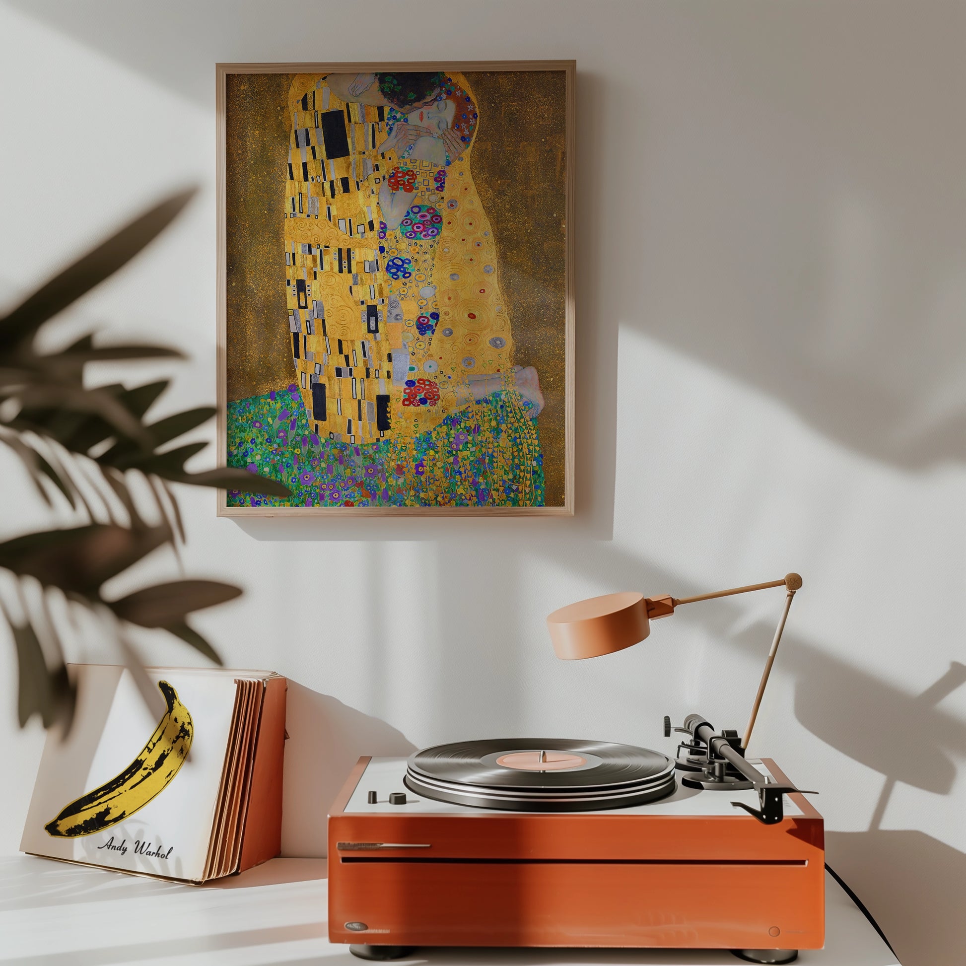 Framed painting of a couple in a golden embrace displayed on a wall above a mid-century record player with an orange finish, surrounded by a potted plant and a stack of album covers.
