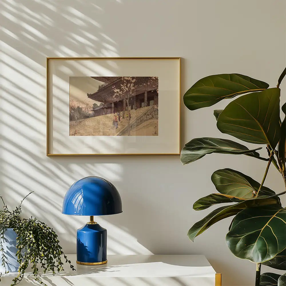 Framed painting of a serene landscape with cherry blossom trees, displayed on a wall near a blue lamp and green plants.