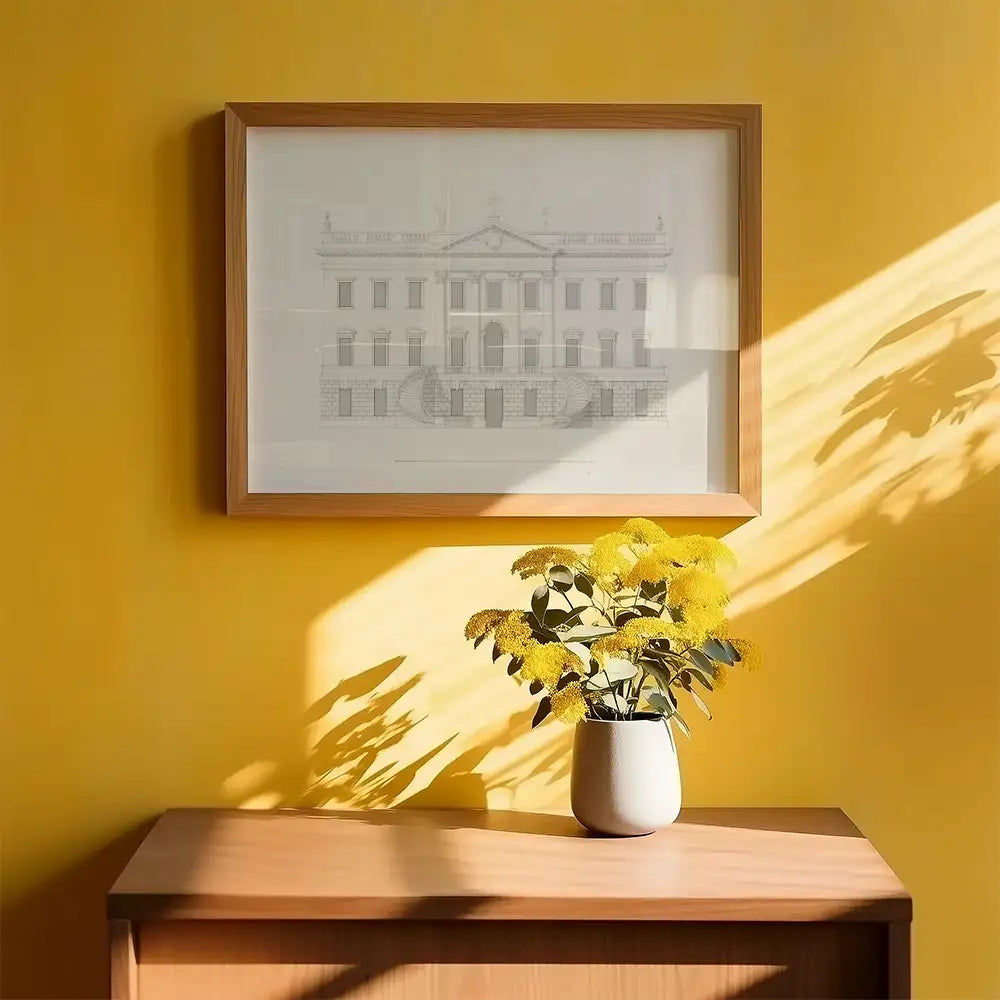 Framed architectural sketch artwork hanging on a yellow wall, with a potted yellow flowering plant on a wooden table.
