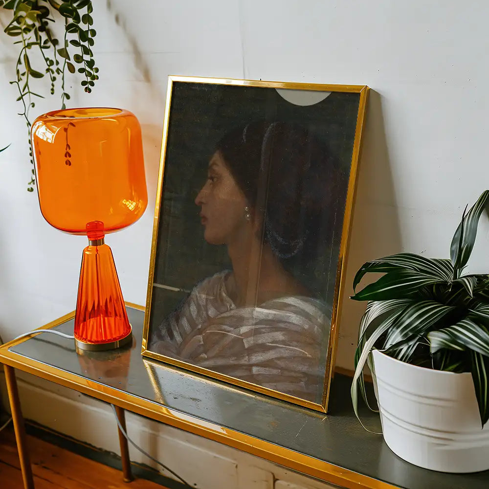 A framed painting of a woman in profile, displayed on a wooden table beside an orange lamp and a potted plant.