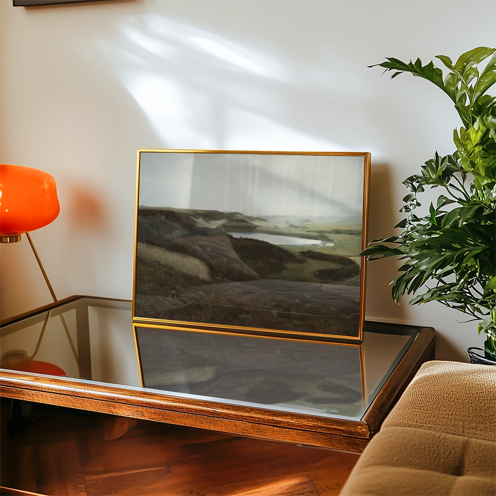 Landscape painting in a gold frame displayed on a coffee table with a modern lamp and potted plant in the background.