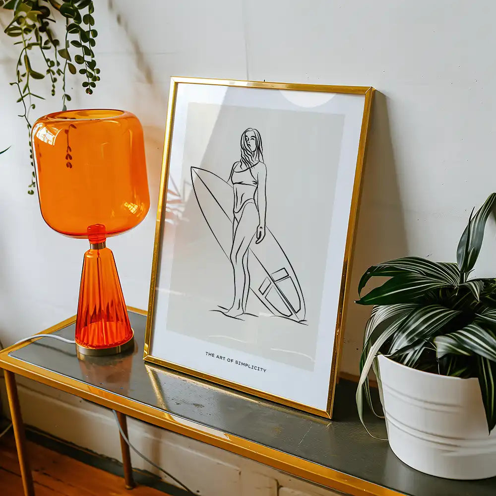 Modern minimalist poster of a woman with a surfboard titled "The Art of Simplicity," framed in gold, displayed alongside an orange lamp and indoor plants.