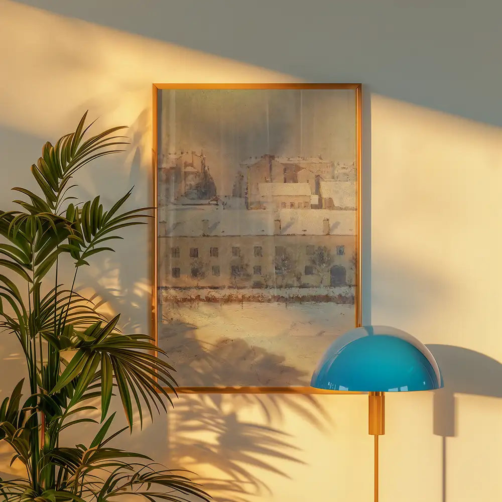 Framed painting of a winter landscape with buildings, set against a wall with shadows from a nearby plant and a lamp with a blue shade.