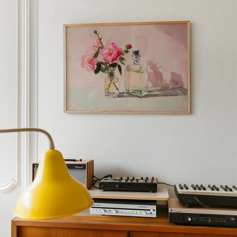 Painting of pink roses in a glass vase, set against a soft pink background, framed and displayed above a wooden shelf with a yellow lamp and music equipment.