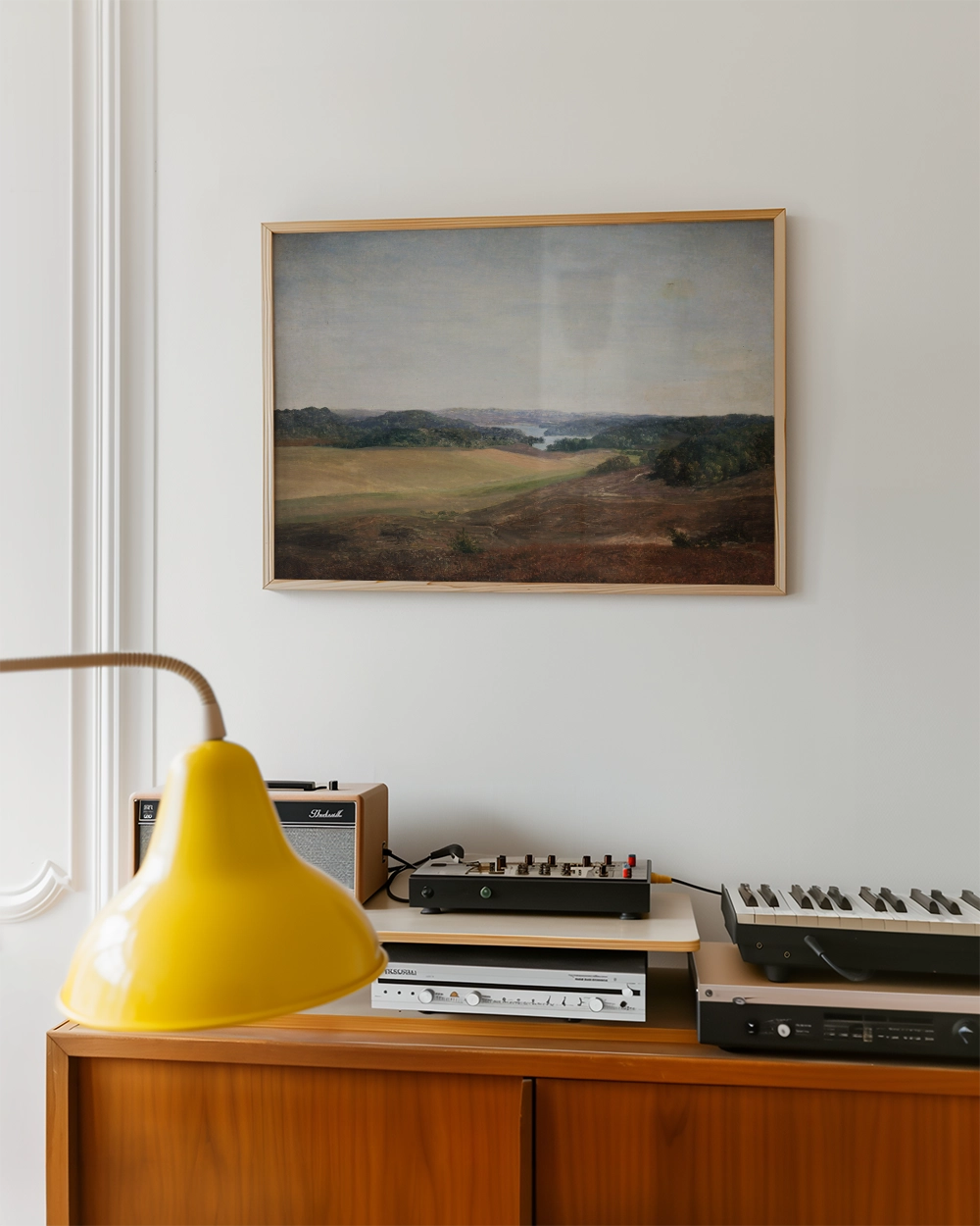 Framed landscape painting hanging on a wall above a wooden cabinet with a yellow lamp and audio equipment.