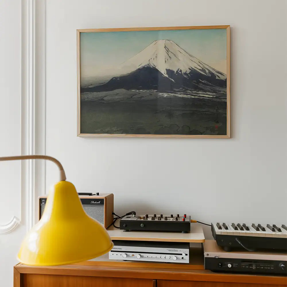 Painting of a mountain with snow-capped peak framed and displayed on a wall above a wooden console table featuring a yellow lamp and audio equipment.