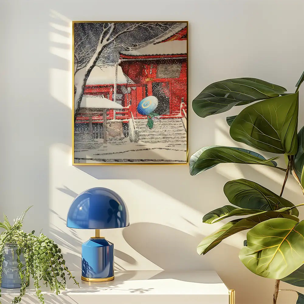 Framed painting of a snow-covered house with red walls and a blue umbrella, displayed above a blue lamp and surrounded by greenery.
