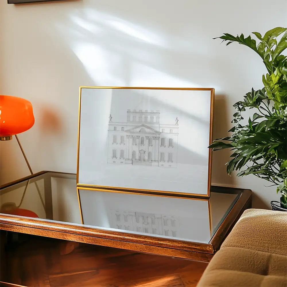 Framed architectural sketch of a historic building displayed on a coffee table with a plant and an orange lamp in a cozy living room setting.
