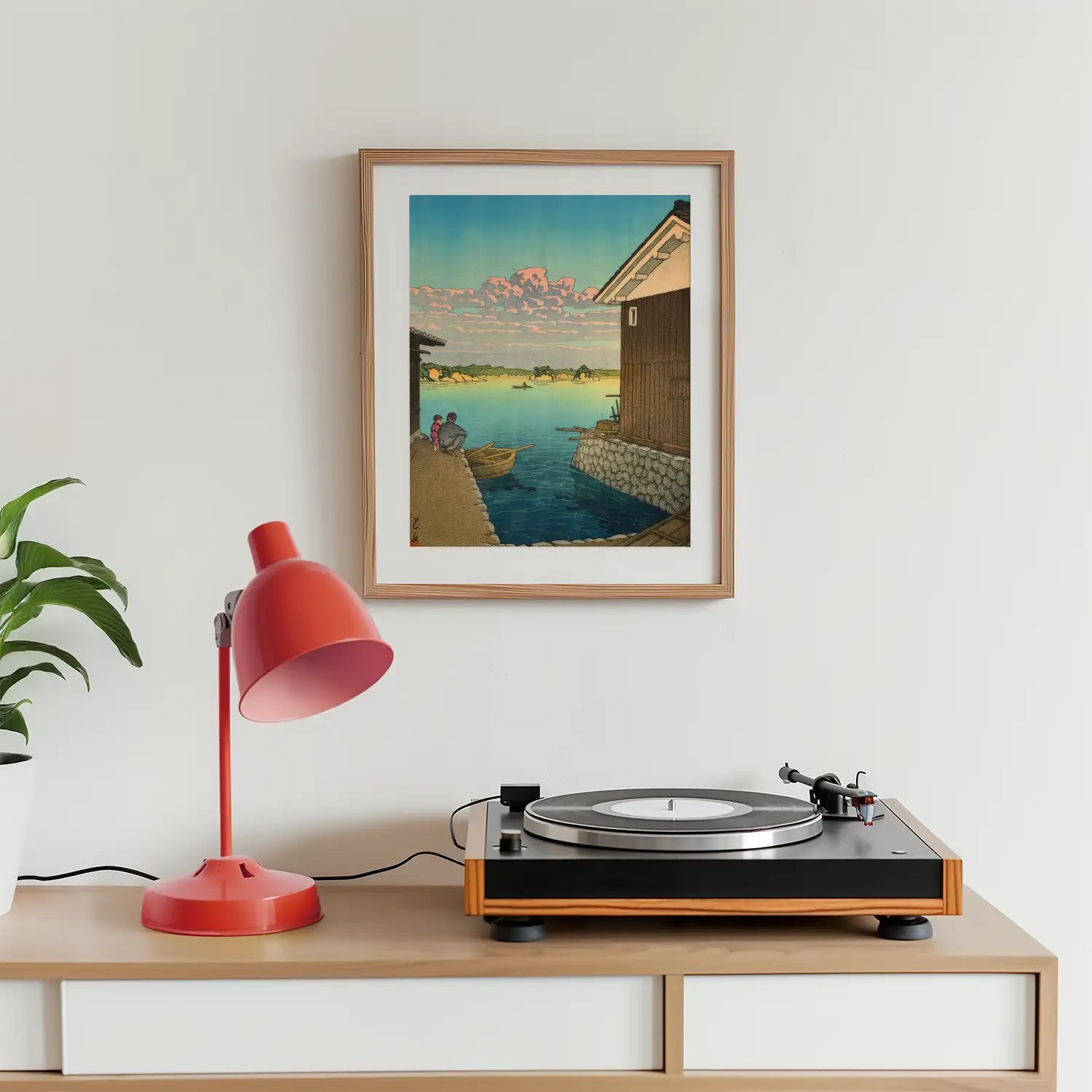 Framed painting of a serene waterfront scene with two figures in a boat, displayed above a record player and a red desk lamp on a wooden surface.