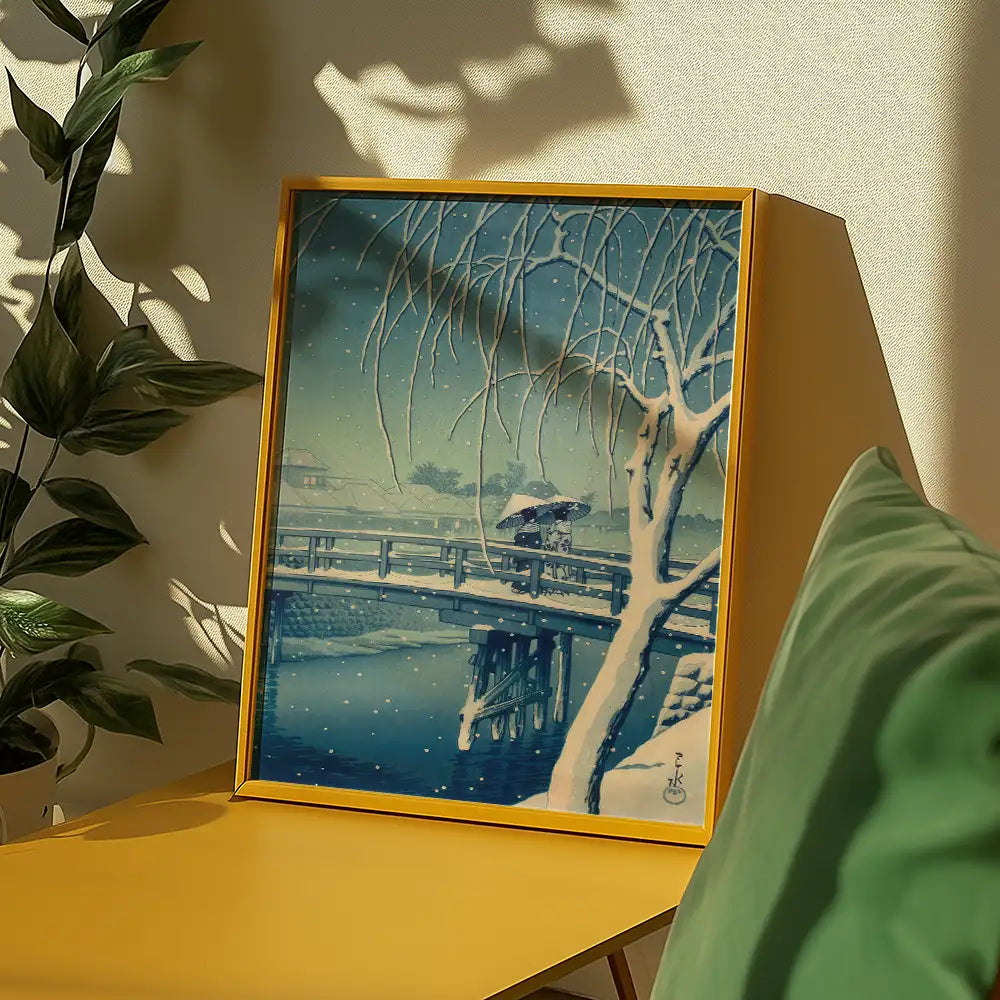 Winter landscape painting featuring a bridge, snow, and a figure with an umbrella, framed in yellow, displayed on a modern yellow table with green cushions and indoor plants in the background.