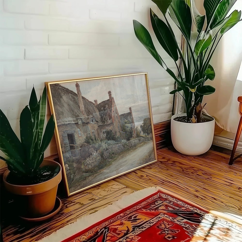 Painting of a rustic cottage landscape framed in gold, positioned beside a potted plant in a cozy interior setting.