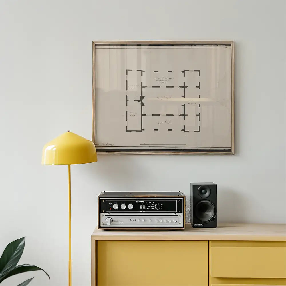 Framed architectural blueprint artwork above a yellow lamp and audio equipment on a wooden cabinet.