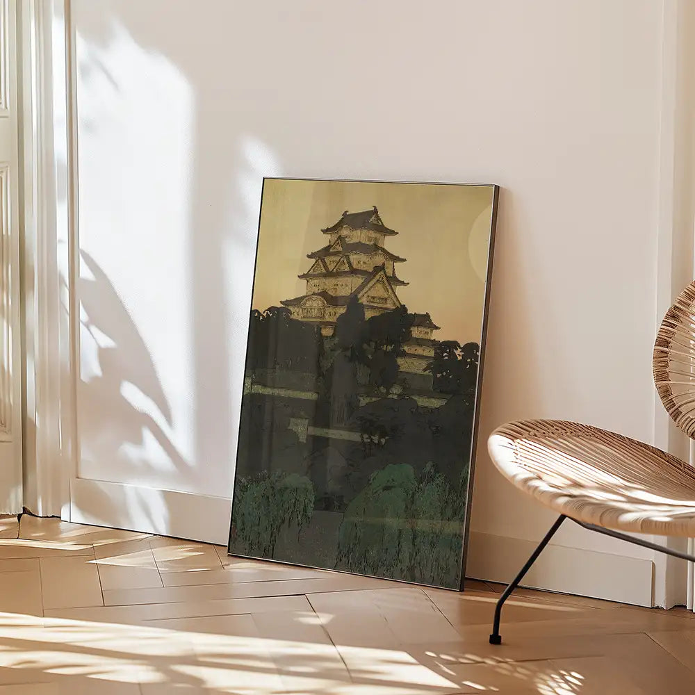 Framed painting of a traditional Japanese castle, displayed against a wall with shadows and a modern chair nearby.