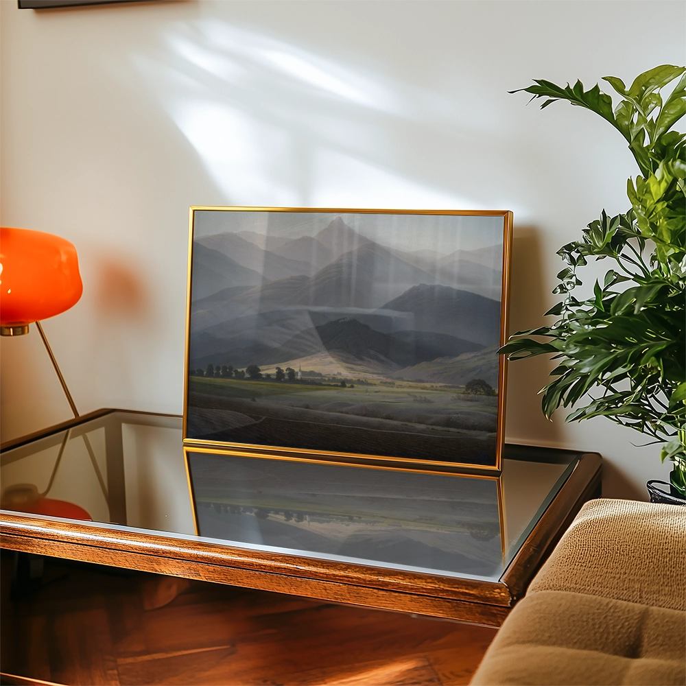 Mountain landscape painting in a gold frame displayed on a glass table with a plant and an orange lamp in the background.