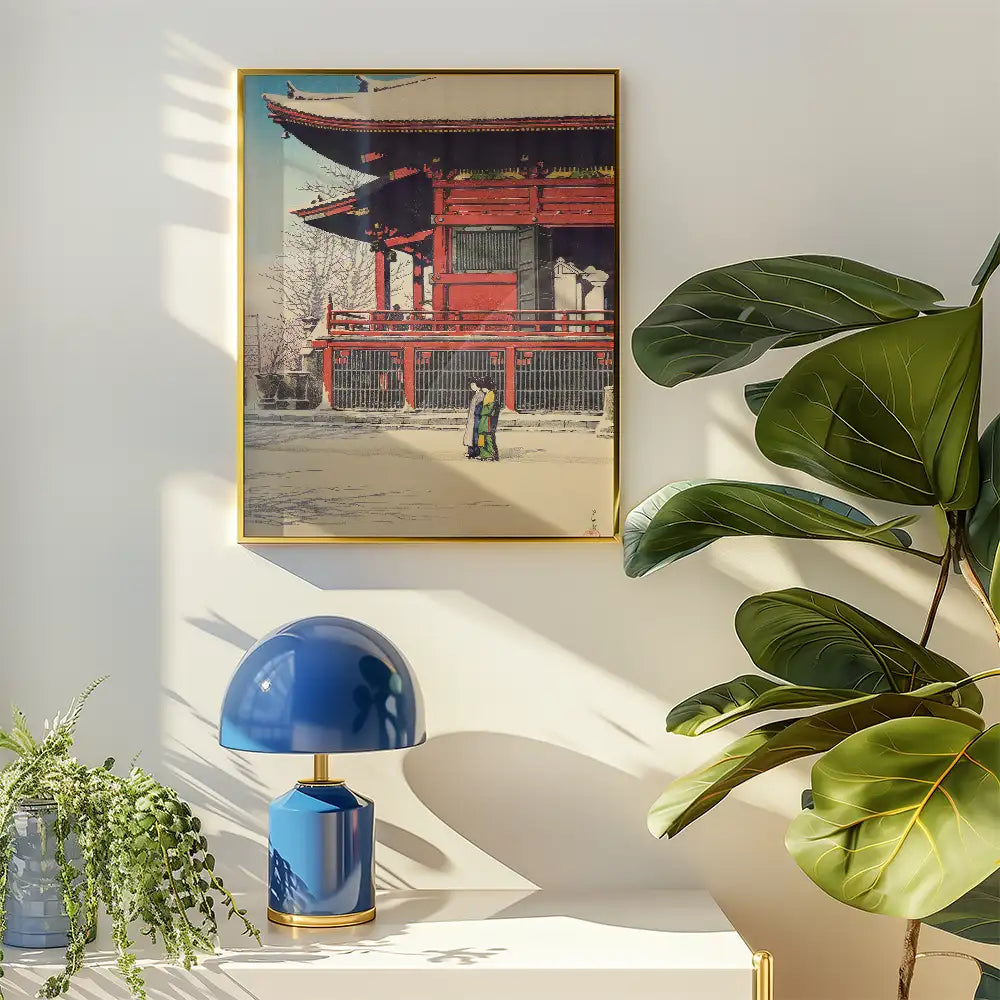 Framed painting of a traditional building with two figures standing in front, displayed on a wall next to a blue lamp and green plants.
