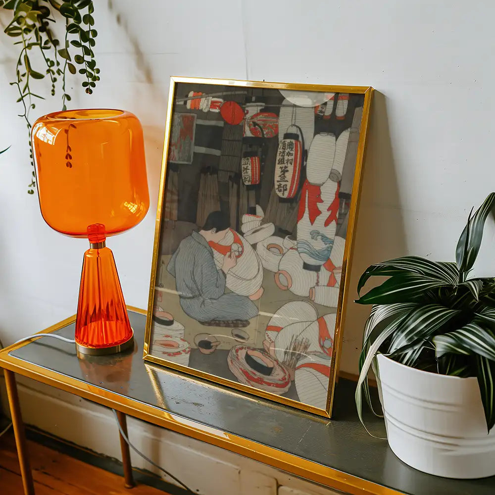 Framed painting of people in a traditional market setting, displayed alongside an orange lamp and a potted plant on a modern table.