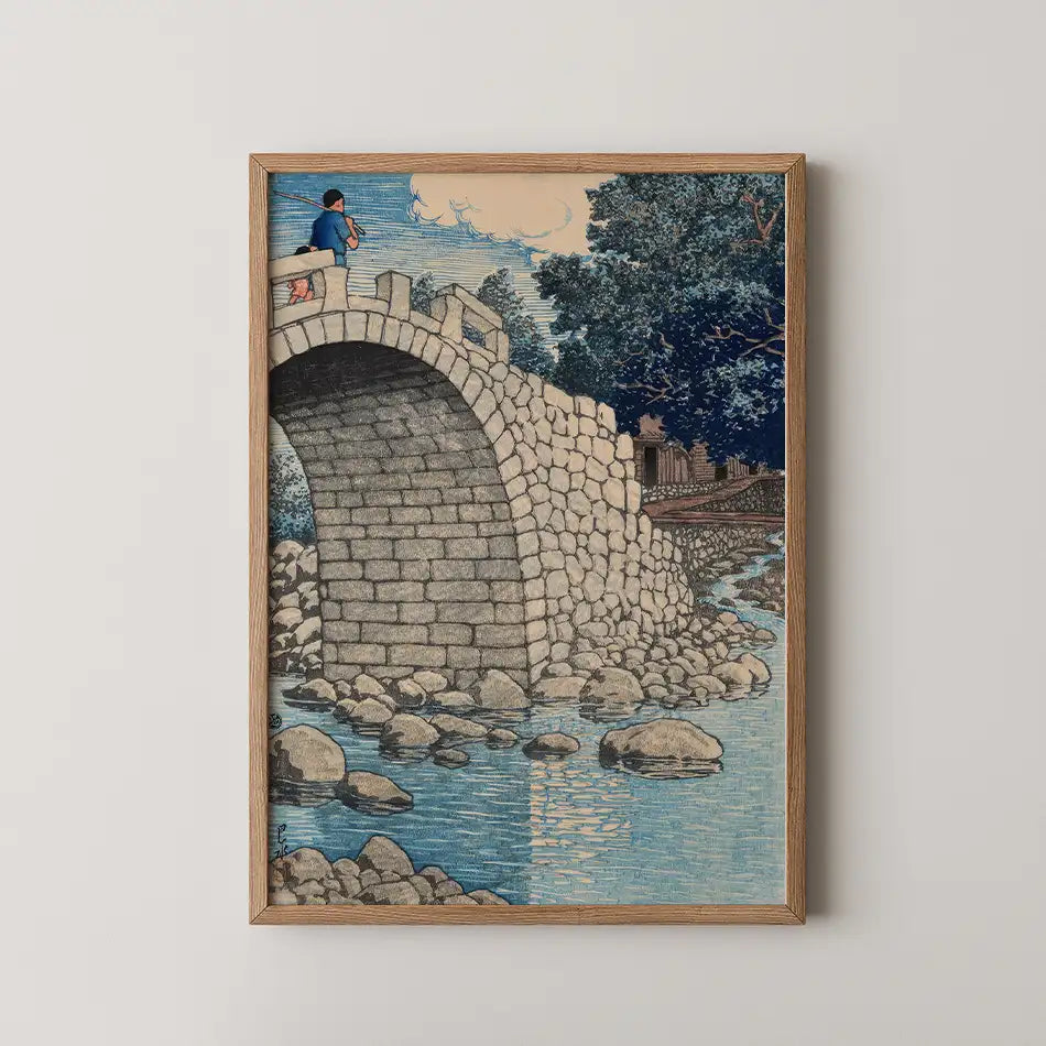 Framed artwork depicting a stone bridge with a person fishing on top, surrounded by a tranquil river and lush greenery.