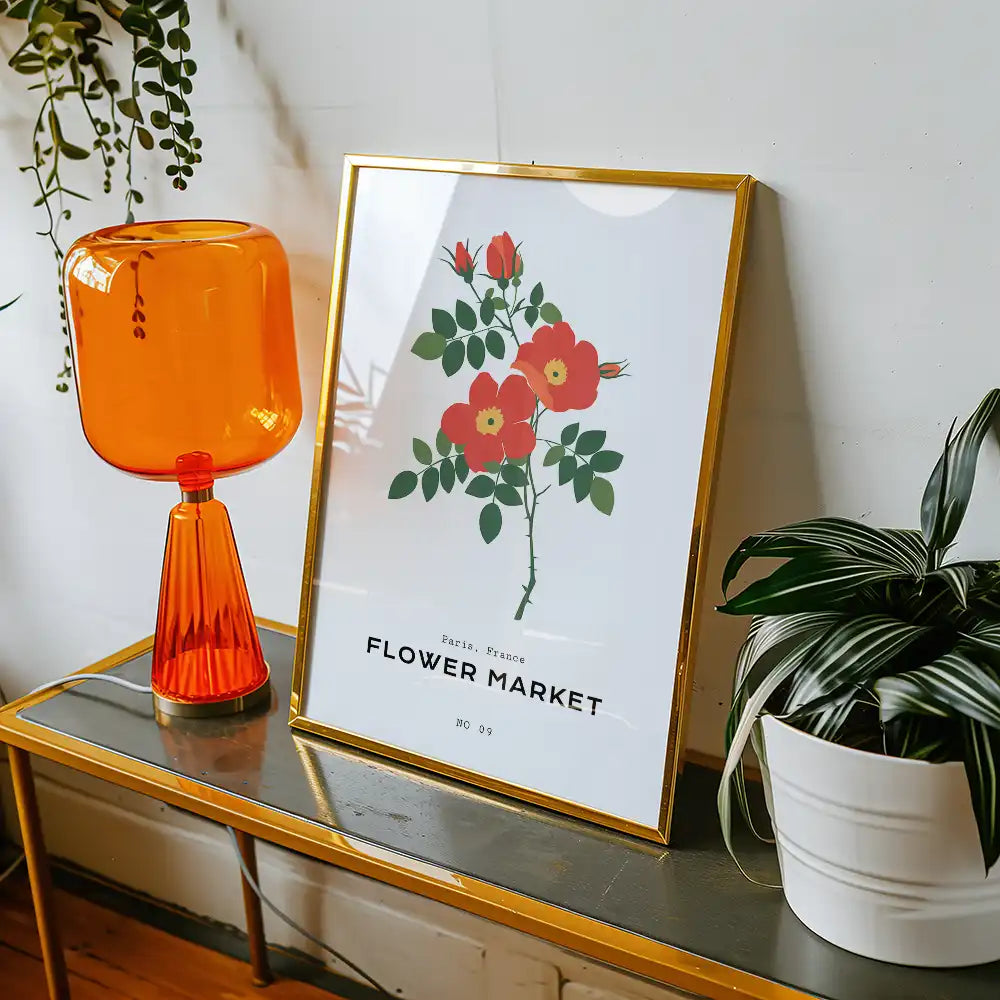 Modern floral poster titled "Flower Market" featuring vibrant red flowers and green leaves, stylishly framed in gold, displayed on a table with an orange lamp and indoor plant.