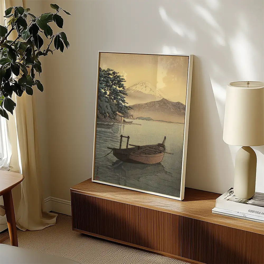 A framed painting of a serene lake scene featuring a small boat, mountains, and trees, displayed on a wooden cabinet next to a lamp in a cozy room.