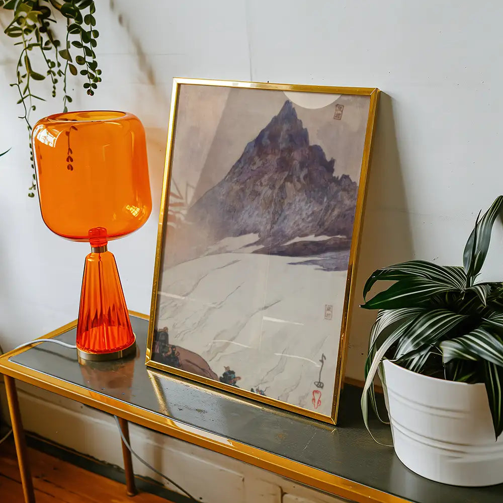 Framed mountain painting displayed on a modern console table beside an orange lamp and a decorative plant.