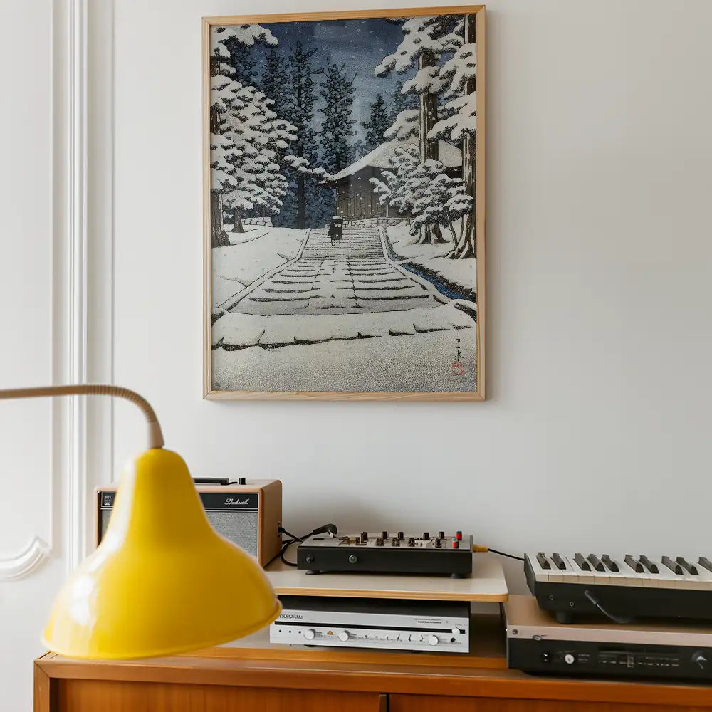 Painting of a snowy landscape with a house and trees, framed and displayed above a wooden console with vintage audio equipment and a yellow lamp.