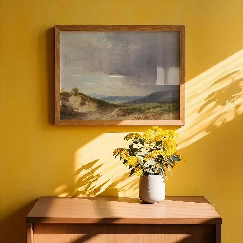 Framed landscape painting featuring rolling hills and a cloudy sky, hanging on a bright yellow wall above a wooden table with a vase of yellow flowers.