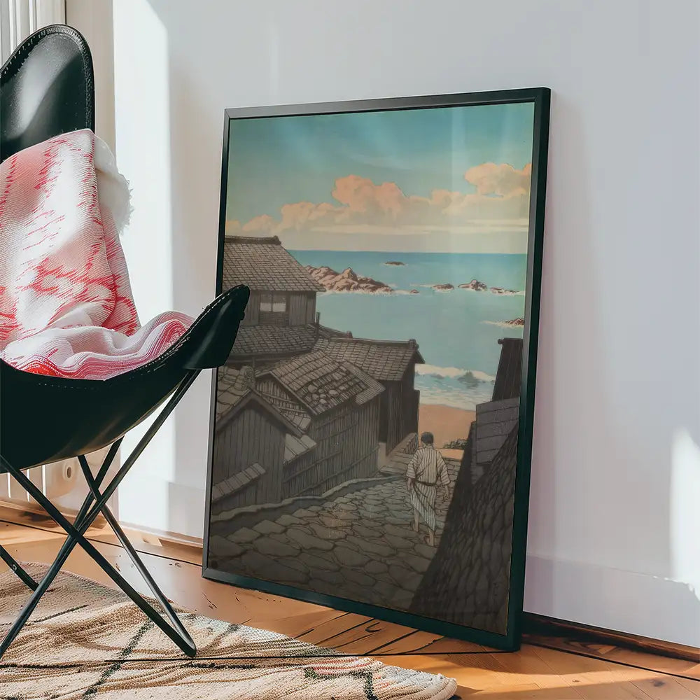 Framed painting of a serene coastal landscape with a person walking down a stone path, leaning against a wall with a black chair and cozy blanket nearby.