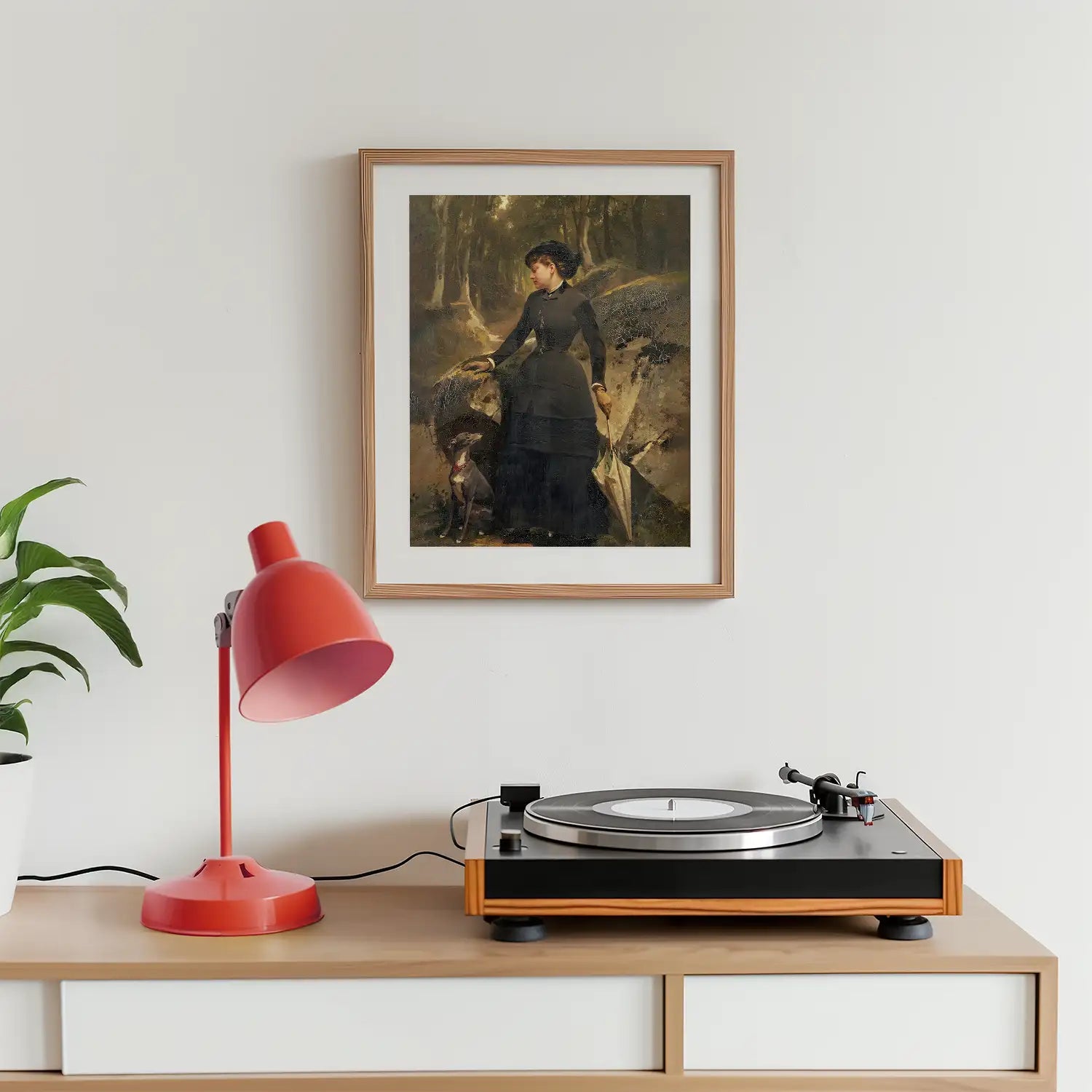 Framed painting of a woman with an umbrella and a dog in a forest, displayed on a wall above a record player and a red desk lamp, with a potted plant nearby.