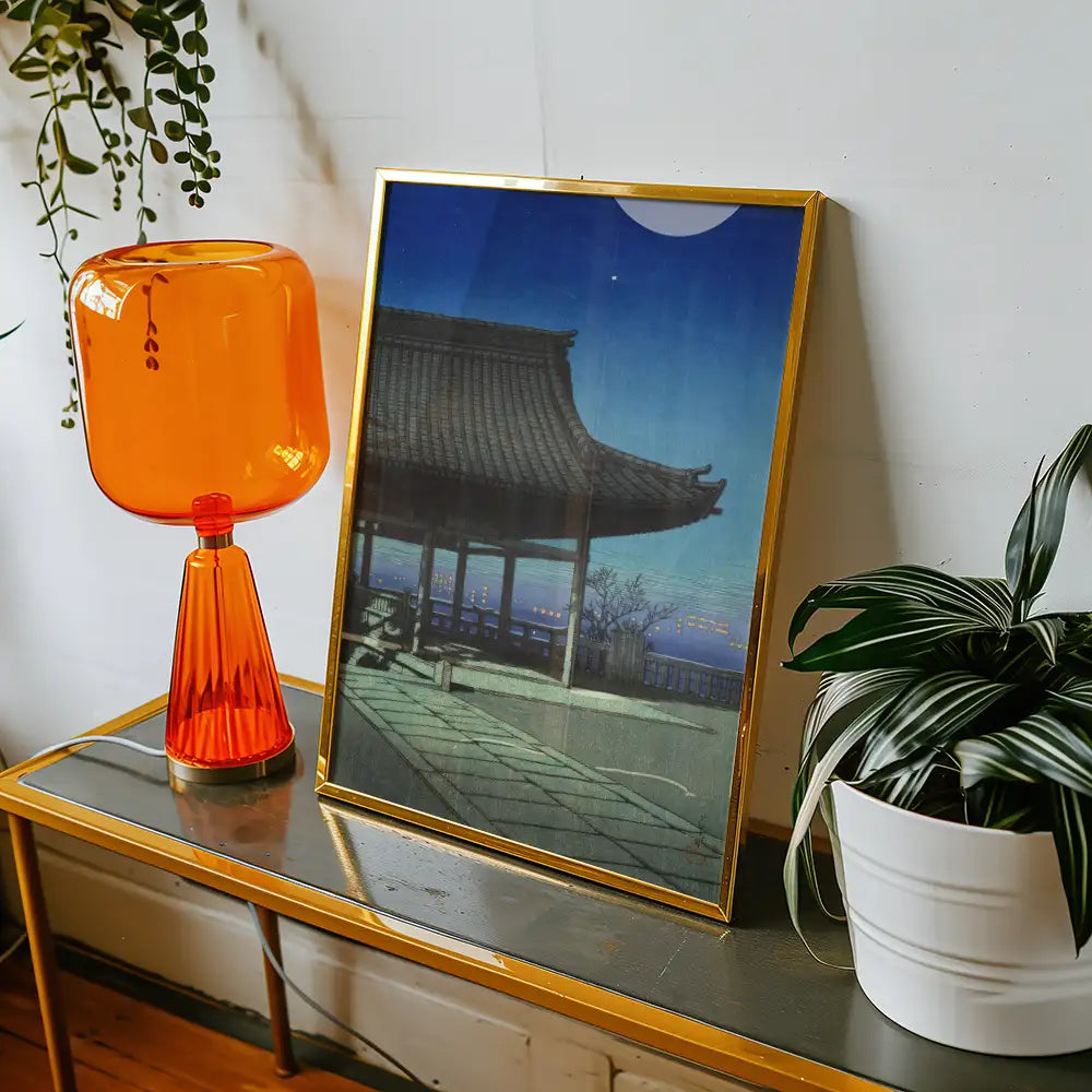 Framed painting of a traditional Asian pavilion with a blue night sky, displayed on a table next to an orange lamp and a potted plant.
