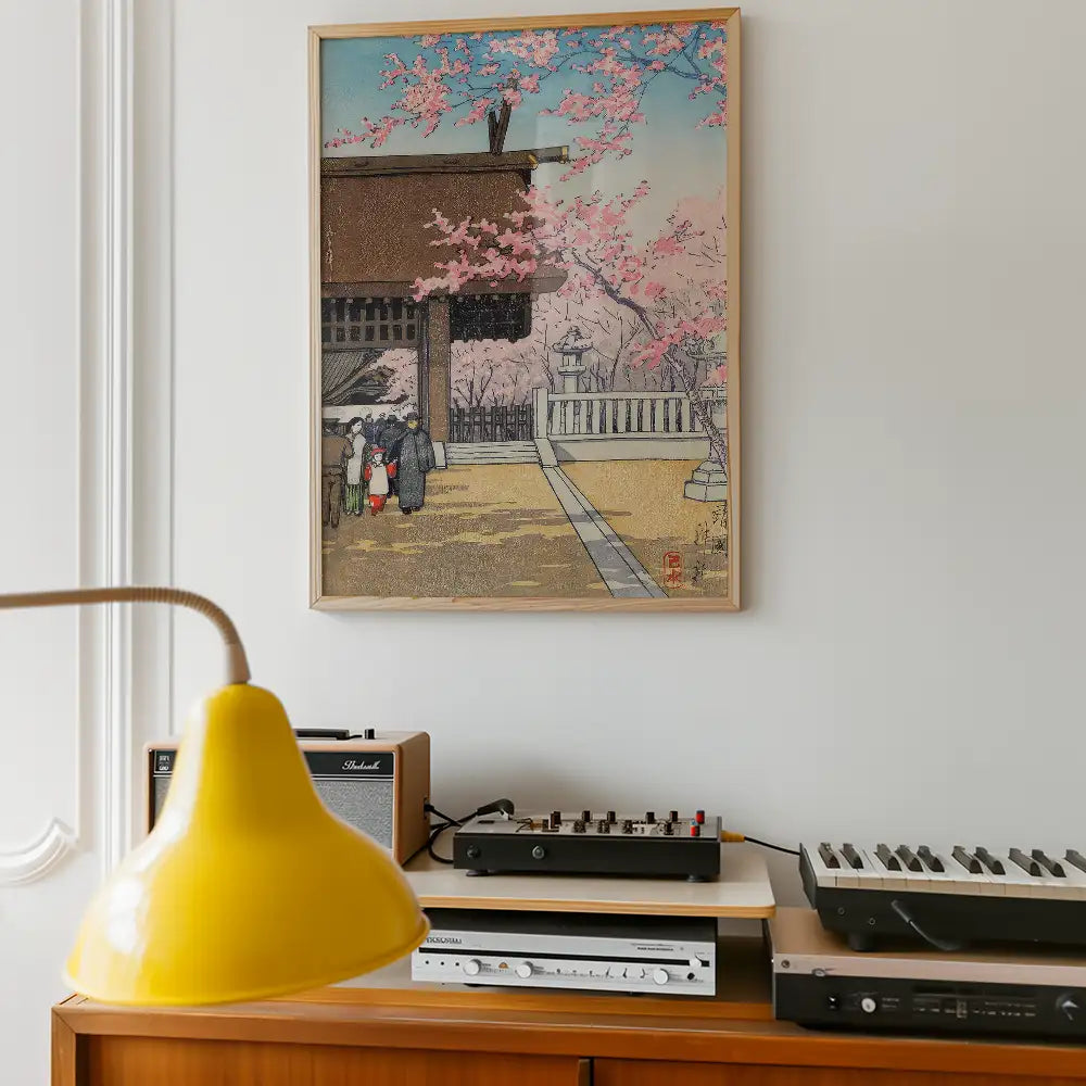 Framed painting of a scenic landscape with cherry blossoms, a traditional building, and people walking, displayed on a wall above a wooden shelf with a yellow lamp and audio equipment.