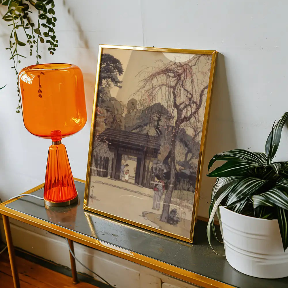 Framed painting of a serene landscape featuring a traditional gate, displayed on a table alongside an orange lamp and a potted plant.