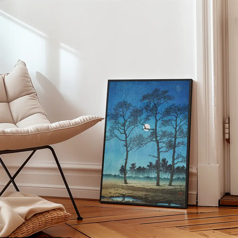 Framed painting of trees against a blue sky, leaning against a wall in a cozy interior setting with a chair and wooden floor.
