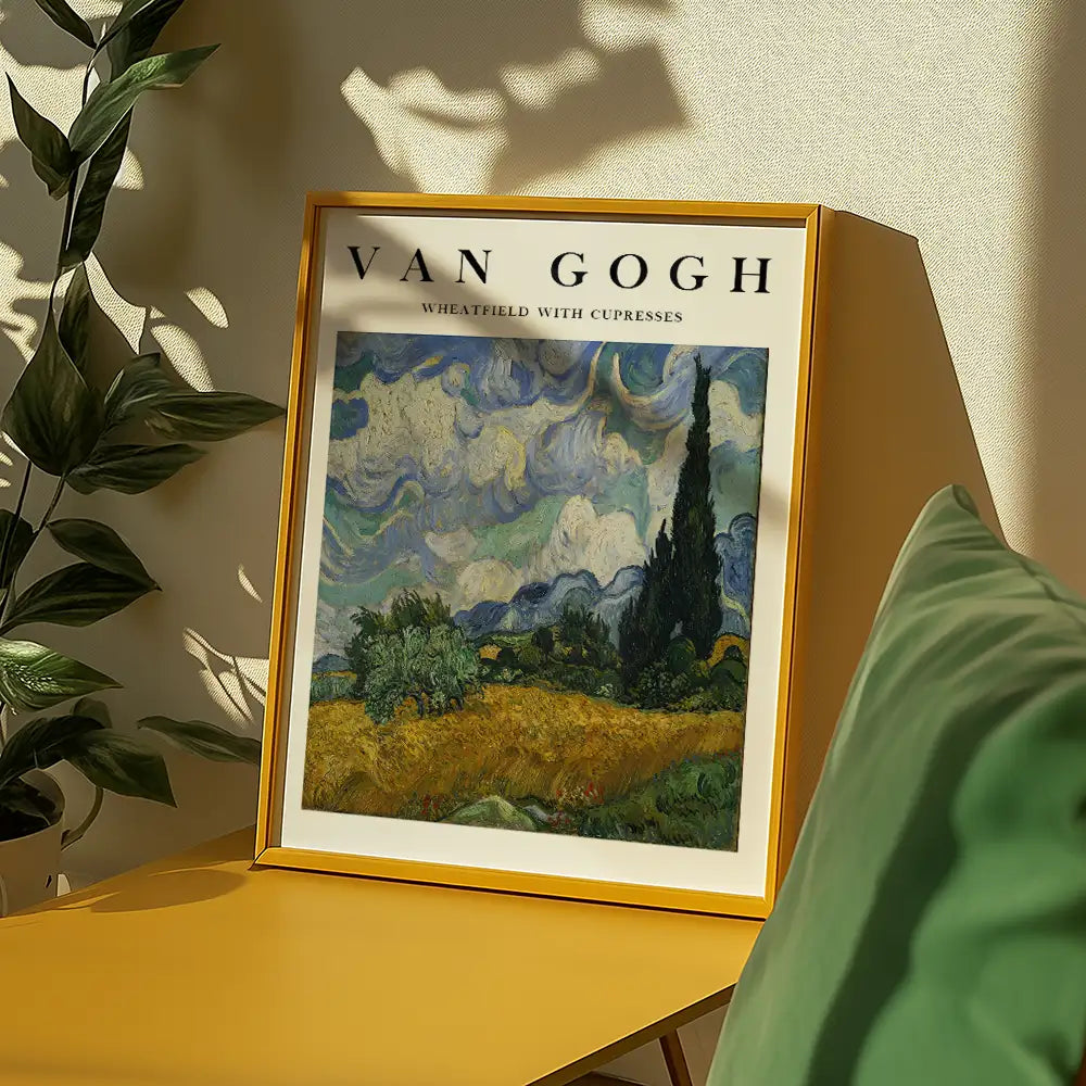 Painting of "Wheatfield with Cypresses" by Vincent van Gogh in a decorative frame, displayed on a yellow table with green plant leaves in the background.