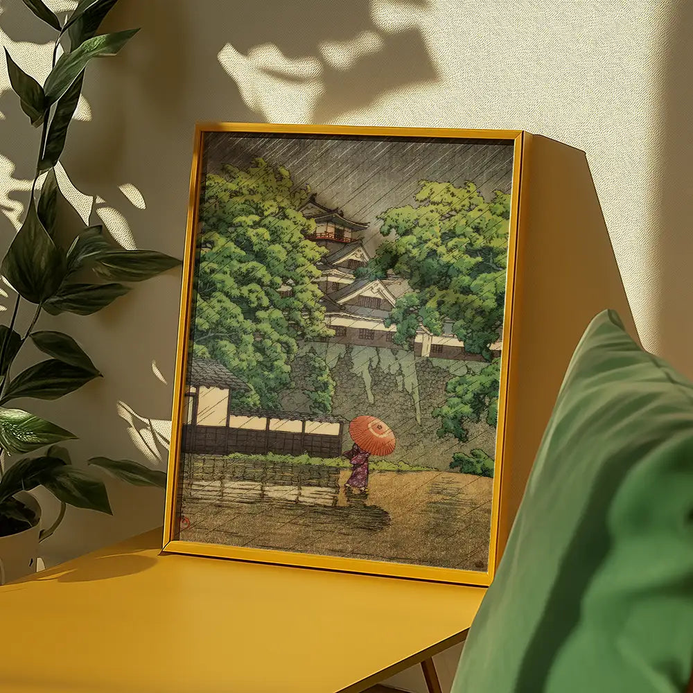 Painting of a woman in a purple kimono walking with an umbrella through a rainy landscape, framed in a yellow frame on a modern table with a green cushion and plant in the background.