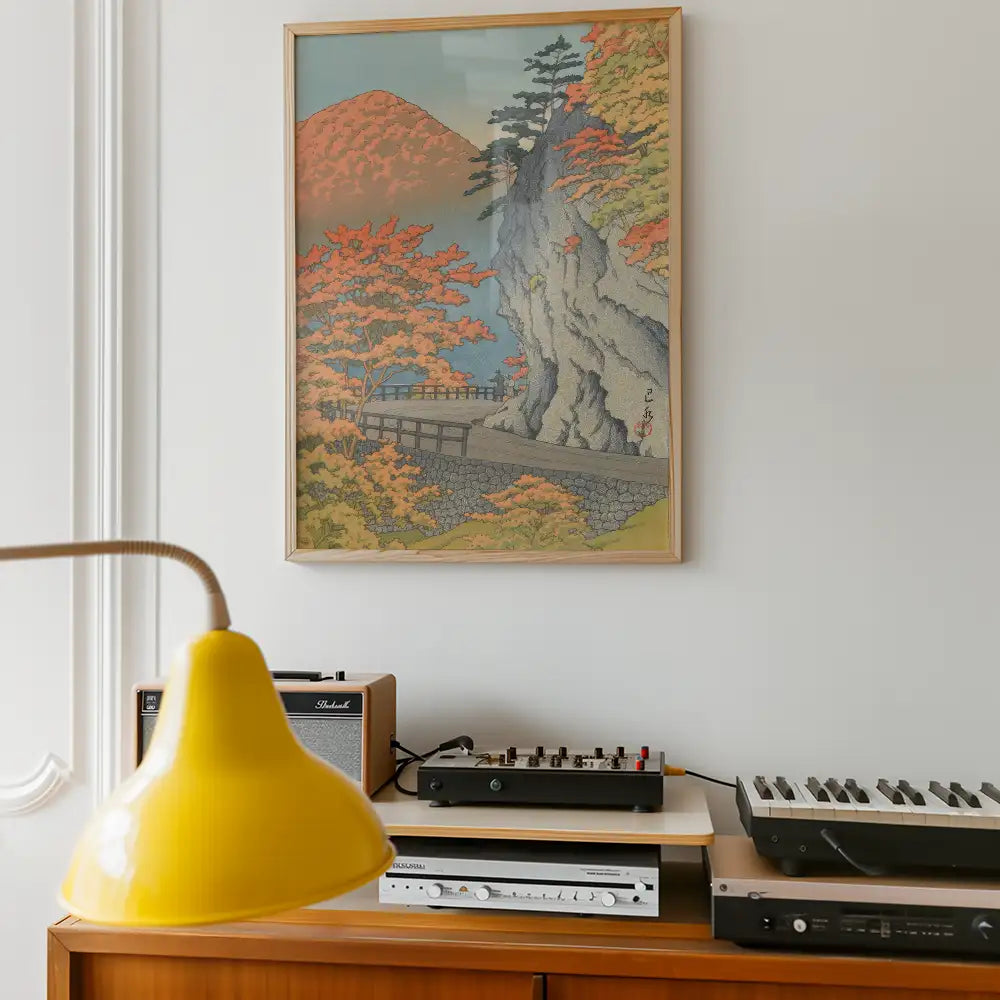 Framed painting of a serene landscape with autumn trees, mountains, and a bridge, displayed on a wall above a wooden cabinet with audio equipment and a yellow lamp.
