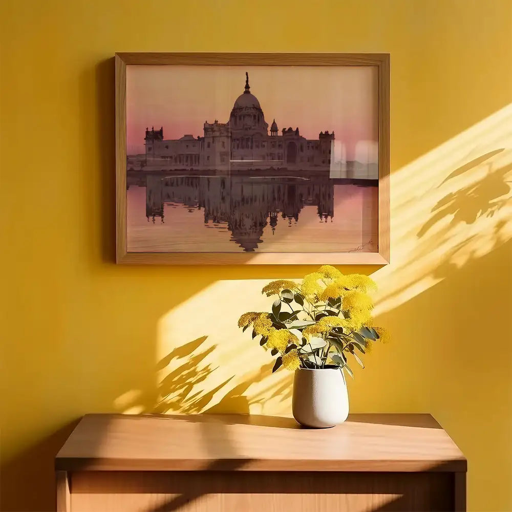 Framed painting of a historic building reflecting in water, displayed on a yellow wall above a wooden dresser with a vase of yellow flowers.