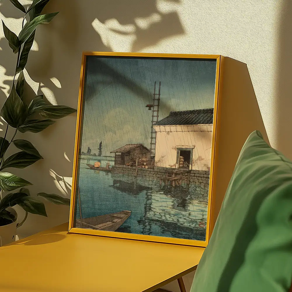 Framed painting of a serene waterside scene featuring a small house and fishing structures, displayed on a yellow surface alongside green decor.